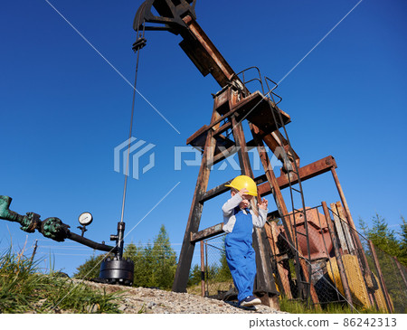 Low angle view of small young power engineer near oil rig. Boy in overalls against the blue sky, petroleum pipeline and pump jack for extracting natural fuel. 86242313