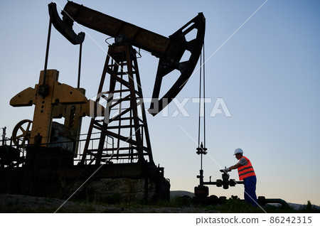 Male mechanic in work clothing checking pump nearby oil derrick. Oil pump operation specialist providing continuous work of drilling rig. Power engineer controlling petroleum pumping station. 86242315