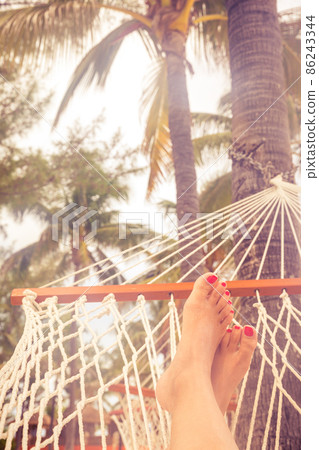 Female legs in a hammock on a background of the sea, palm trees and sunset. Vacation concept with orange color tone 86243344