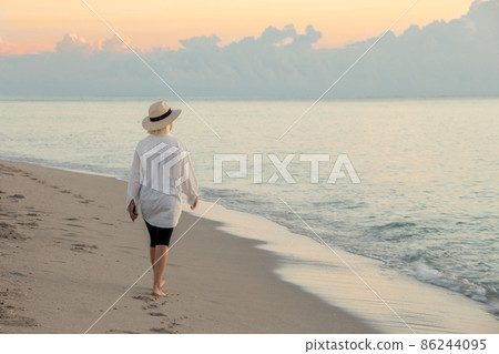 Woman holding cell phone and wearing straw hat walking on a beach at sunset Woman holding cell phone and wearing straw hat walking on a beach at sunset 86244095