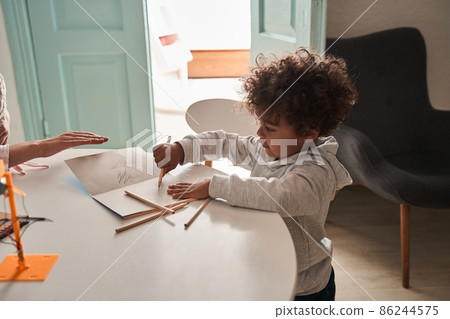Brunette curly boy standing at the table and drawing while spending time at the kitchen Brunette curly boy standing at the table and drawing while spending time at the kitchen 86244575