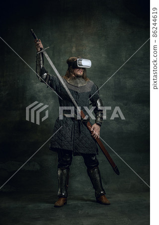 Portrait of man, medieval warrior, knight with dirty wounded face in VR headset isolated over dark vintage background. Comparison of eras, history Portrait of man, medieval warrior, knight with dirty wounded face in VR headset isolated over dark vintage background. Comparison of eras, history 86244619