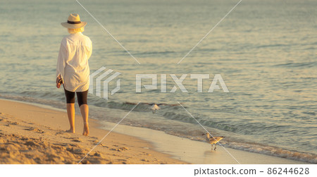 Woman wearing white shirt and straw hat walking on beach at sunset Woman wearing white shirt and straw hat walking on beach at sunset 86244628