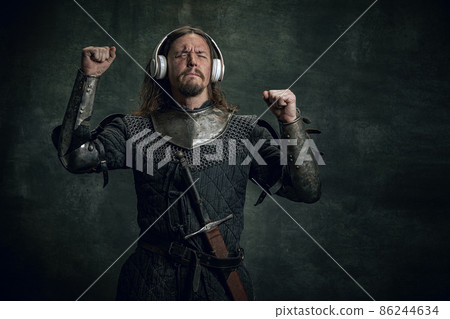 Young medieval warrior or knight with dirty wounded face in headphones listening to music isolated over dark vintage background. Comparison of eras, history 86244634