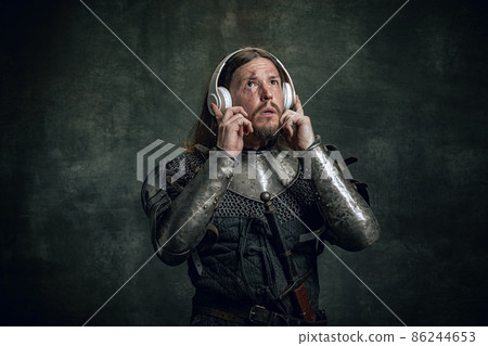 Shocked medieval warrior or knight with dirty wounded face in headphones listening to music isolated over dark vintage background. Comparison of eras, history 86244653