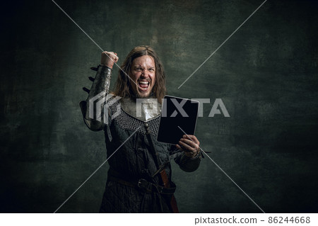 Vintage style portrait of brutal smiling man, medieval warrior or knight with dirty wounded face using modern gadget isolated over dark background. Comparison of eras Vintage style portrait of brutal smiling man, medieval warrior or knight with dirty wounded face using modern gadget isolated over dark background. Comparison of eras 86244668