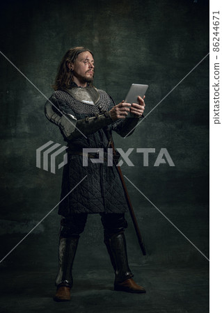 Vintage style portrait of brutal smiling man, medieval warrior or knight with dirty wounded face using modern gadget isolated over dark background. Comparison of eras Vintage style portrait of brutal smiling man, medieval warrior or knight with dirty wounded face using modern gadget isolated over dark background. Comparison of eras 86244671