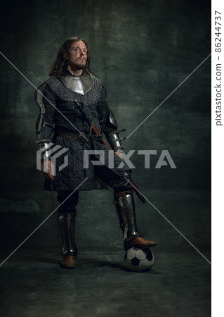 Portrait of medieval warrior or knight with dirty wounded face posing with football ball isolated over dark background. Comparison of eras, history 86244737