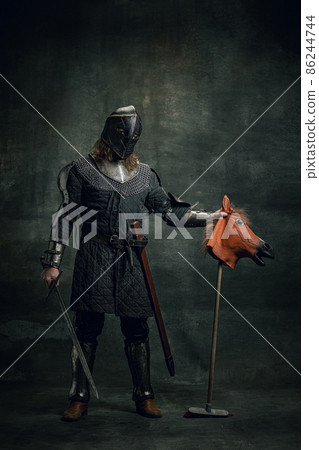 Portrait of medieval warrior or knight wearing helmet and armor riding toy horse, holding big sword isolated over dark background. Comparison of eras, history 86244744
