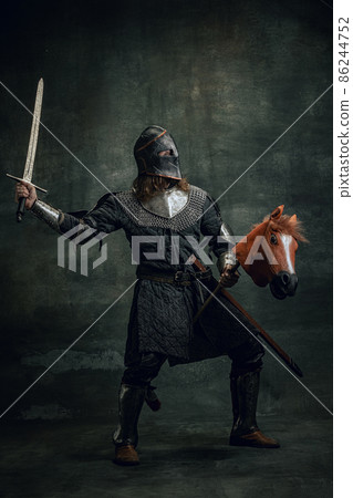 Portrait of medieval warrior or knight wearing helmet and armor riding toy horse, holding big sword isolated over dark background. Comparison of eras, history 86244752