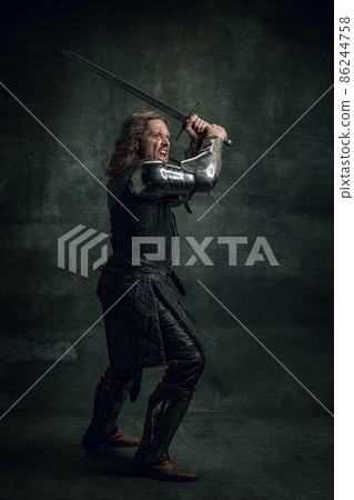 Vintage style portrait of brutal seriuos man, medieval warrior or knight with dirty wounded face holding big sword isolated over dark background. Comparison of eras 86244758