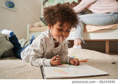 Little multiracial boy laying at the floor and drawing something with pencils at the paper Little multiracial boy laying at the floor and drawing something with pencils at the paper 86244846