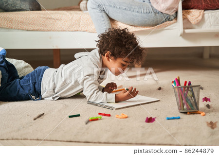 Little multiracial boy laying at the floor and drawing something with pencils at the paper Little multiracial boy laying at the floor and drawing something with pencils at the paper 86244879