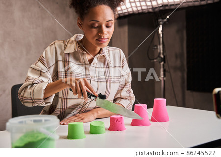 Woman looking gently and holding knife while cutting kinetic sand during recording video blog Woman looking gently and holding knife while cutting kinetic sand during recording video blog 86245522