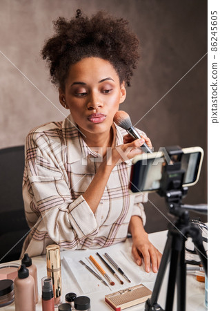 Attentive curly multiracial woman applying makeup on her face while streaming 86245605