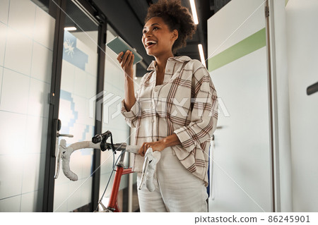 Joyful multiracial woman standing with bike at the hall and recording voice message Joyful multiracial woman standing with bike at the hall and recording voice message 86245901