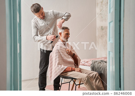 Elderly handsome man getting his hair and beard cut at home while his careful son helping Elderly handsome man getting his hair and beard cut at home while his careful son helping 86246008