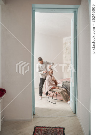 Adult man cutting his retirement father with trimmer while spending time at home Adult man cutting his retirement father with trimmer while spending time at home 86246009