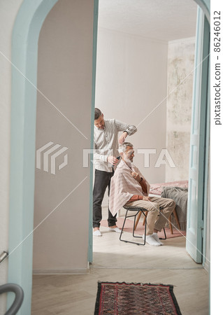 Adult son cutting hair of his senior father while he sitting at the modern flat calmly Adult son cutting hair of his senior father while he sitting at the modern flat calmly 86246012