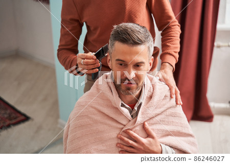 Man wearing towel sitting at the chair while his father making haircut for him with the trimmer Man wearing towel sitting at the chair while his father making haircut for him with the trimmer 86246027