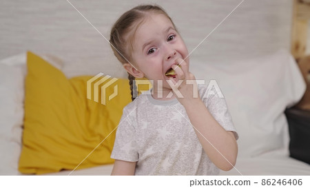 little girl eating potato chips while sitting on the bed, enjoying taste of crisp at home, useless food with spices, seasoned food snack small child, happiness in children's variety goodies vegetable 86246406