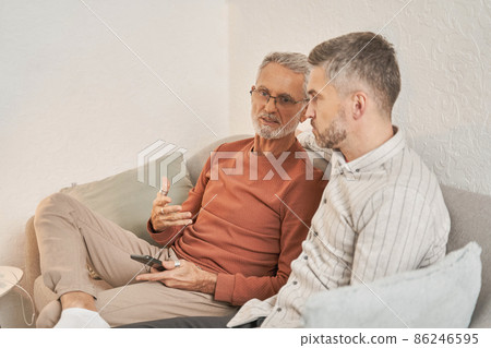 Smiling senior father sitting on couch and talking with his son while spending time at home Smiling senior father sitting on couch and talking with his son while spending time at home 86246595