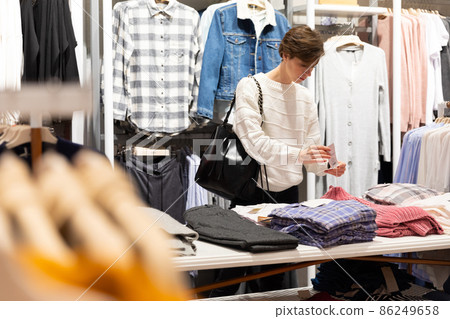 Young beautiful brunette woman with a short haircut in a white sweater chooses stylish clothes in a Young beautiful brunette woman with a short haircut in a white sweater chooses stylish clothes in a 86249658