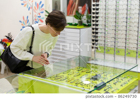 Photo of young attractive brunet woman with short haircut in a casual white sweater chooses glasses 86249730