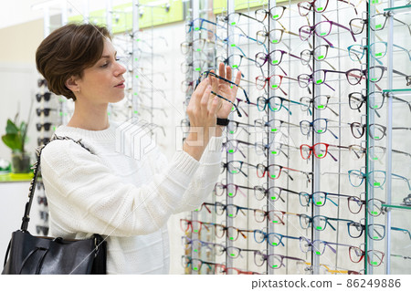 Photo of young thinking attractive brunette woman in a white sweater chooses glasses for vision on a Photo of young thinking attractive brunette woman in a white sweater chooses glasses for vision on a 86249886
