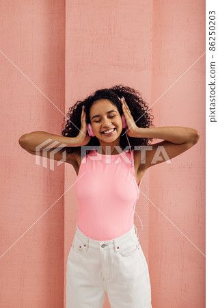 Smiling woman with closed eyes listening to music by pink headphones standing at pink wall 86250203