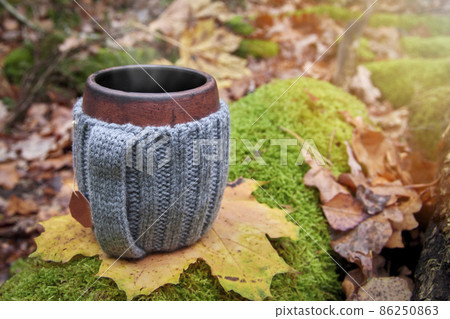 Clay mug in woolen knitted cover. Steam from the mug on the sunny background of autumn forest. Clay mug in woolen knitted cover. Steam from the mug on the sunny background of autumn forest. 86250863