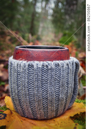 Clay mug in woolen knitted cover. Steam from the mug on the dark background of autumn forest Clay mug in woolen knitted cover. Steam from the mug on the dark background of autumn forest 86250867