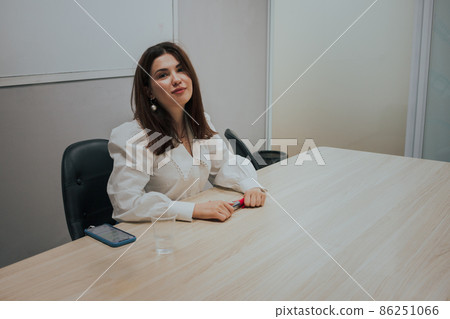 Young brunette woman in a white blouse sitting at the table in the office, holding a marker in her hands and looking at the camera. Business Education concept. Young brunette woman in a white blouse sitting at the table in the office, holding a marker in her hands and looking at the camera. Business Education concept. 86251066