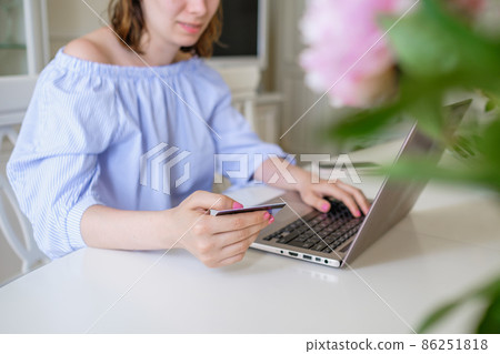 Young woman in stylish blouse makes purchases online via laptop entering bank card details behind flowers on foreground at home closeup 86251818