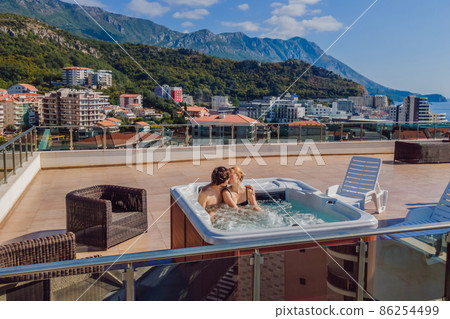 Drone view. Portrait of young carefree happy smiling couple relaxing at hot tub during enjoying happy traveling moment vacation life against the background of green big mountains Drone view. Portrait of young carefree happy smiling couple relaxing at hot tub during enjoying happy traveling moment vacation life against the background of green big mountains 86254499