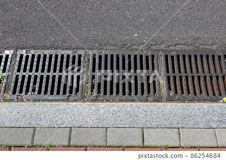 catch basin grate of the lattice of the drain system for drainage of rainwater from the road surface with a tarmac road at granite curb with a stone tile sidewalk close up top view, nobody. catch basin grate of the lattice of the drain system for drainage of rainwater from the road surface with a tarmac road at granite curb with a stone tile sidewalk close up top view, nobody. 86254684