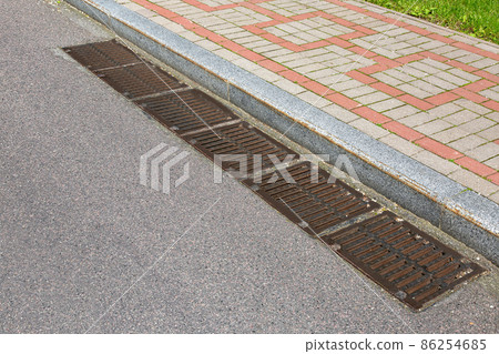 rectangular manholes of the lattice of the drainage system for drainage of rainwater from the road surface with an asphalt road at granite curb with stone tile sidewalk on sidewalk. rectangular manholes of the lattice of the drainage system for drainage of rainwater from the road surface with an asphalt road at granite curb with stone tile sidewalk on sidewalk. 86254685