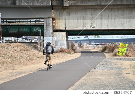 A young man running on a cycling road on a riverbed, a lawn withered in winter and a railway bridge beyond it, a bicycle running in the winter sun 86254767