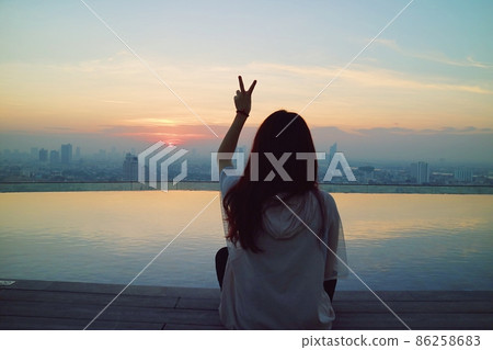 有一個女生坐在頂樓無邊際游泳池前方等待著都市日出的景觀 有一個女生坐在頂樓無邊際游泳池前方等待著都市日出的景觀 86258683