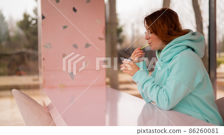 Caucasian red-haired woman eats ice cream with a plastic spoon while sitting alone in a cafe. Caucasian red-haired woman eats ice cream with a plastic spoon while sitting alone in a cafe. 86260081