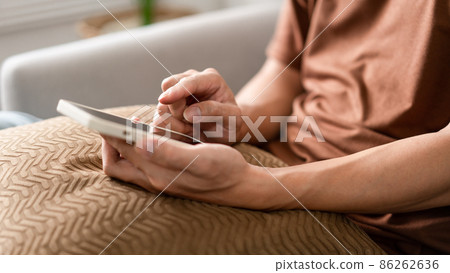 Technology Concept The man sitting next to a pot of plant, putting a small pillow on his lap, and checking something on his smartphone Technology Concept The man sitting next to a pot of plant, putting a small pillow on his lap, and checking something on his smartphone 86262636