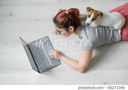 A Caucasian red-haired woman is lying on the floor with her small dog and is typing on a laptop. Female freelancer is working from home in quarantine. A loyal puppy Jack Russell Terrier. 86271366