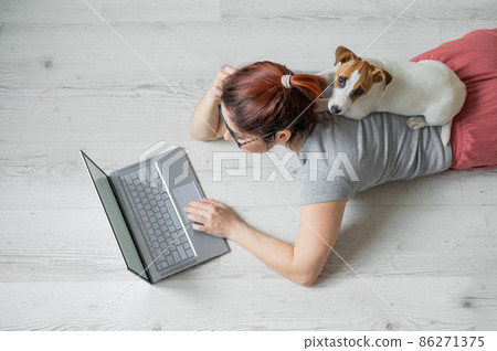 A European red-haired woman is lying on the floor with her dog and is typing on a laptop. Female student studying remotely at home in quarantine. A loyal puppy Jack Russell Terrier. Exam preparation. 86271375