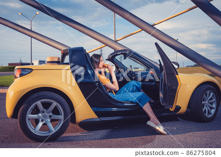Young girl in blue jeans, orange top and flip flops is eating hamburger while sitting in yellow car cabrio. Fast food, unhealthy nutrition Young girl in blue jeans, orange top and flip flops is eating hamburger while sitting in yellow car cabrio. Fast food, unhealthy nutrition 86275804