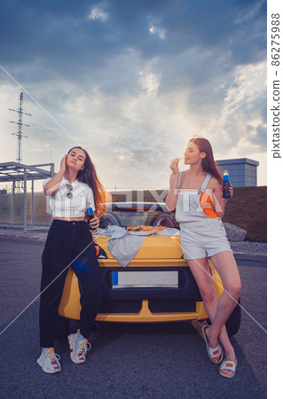Women enjoying soda in glass bottles and eating french fries, leaning on trunk of yellow car with pizza on it. Fast food. Full length, mock up Women enjoying soda in glass bottles and eating french fries, leaning on trunk of yellow car with pizza on it. Fast food. Full length, mock up 86275988