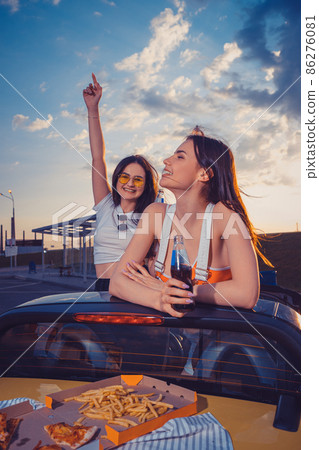 Happy females having fun and drinking soda in glass bottles, posing in yellow car with french fries and pizza on its trunk. Fast food. Copy space Happy females having fun and drinking soda in glass bottles, posing in yellow car with french fries and pizza on its trunk. Fast food. Copy space 86276081