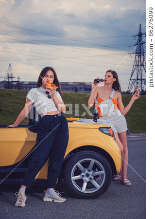 Attractive models in casual outfit eating pizza, enjoying soda water in glass bottles, posing near yellow car on parking lot. Full length, copy space 86276099