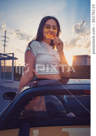 Young female in sunglasses and white t-shirt is eating pizza while posing standing in yellow car cabriolet. Fast food. Summer sunset. Close up 86276119