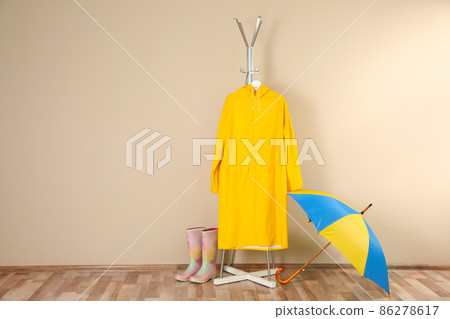 Umbrella, rain coat and rubber boots near beige wall. Space for text 86278617