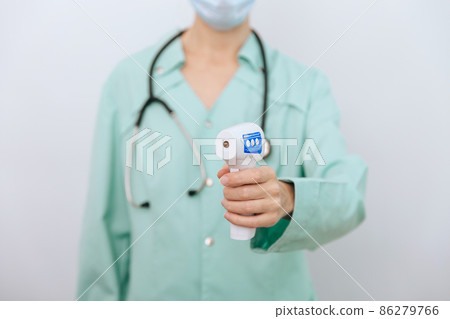 Temperature measurement gun in doctor hands. Close-up shot of doctor wearing protective surgical mask ready to use infrared isometric thermometer gun to check body temperature for virus symptoms. Temperature measurement gun in doctor hands. Close-up shot of doctor wearing protective surgical mask ready to use infrared isometric thermometer gun to check body temperature for virus symptoms. 86279766